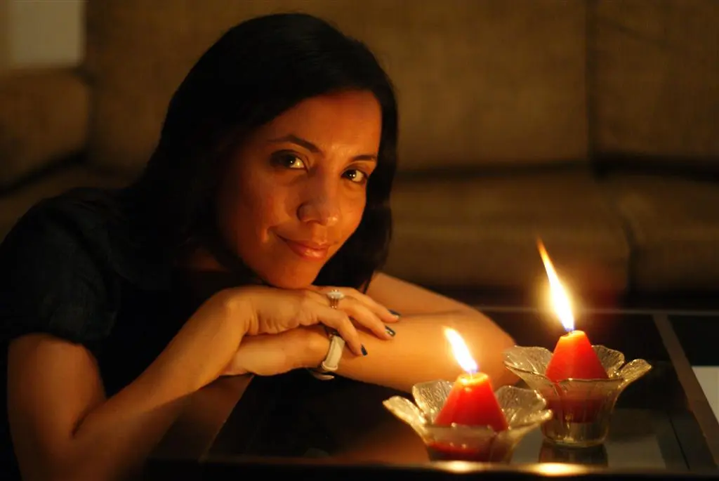 Num lugar bem escuro a luz de duas velas, uma mulher de cabelos compridos e olhos pretos te encara com um leve sorriso no rosto.